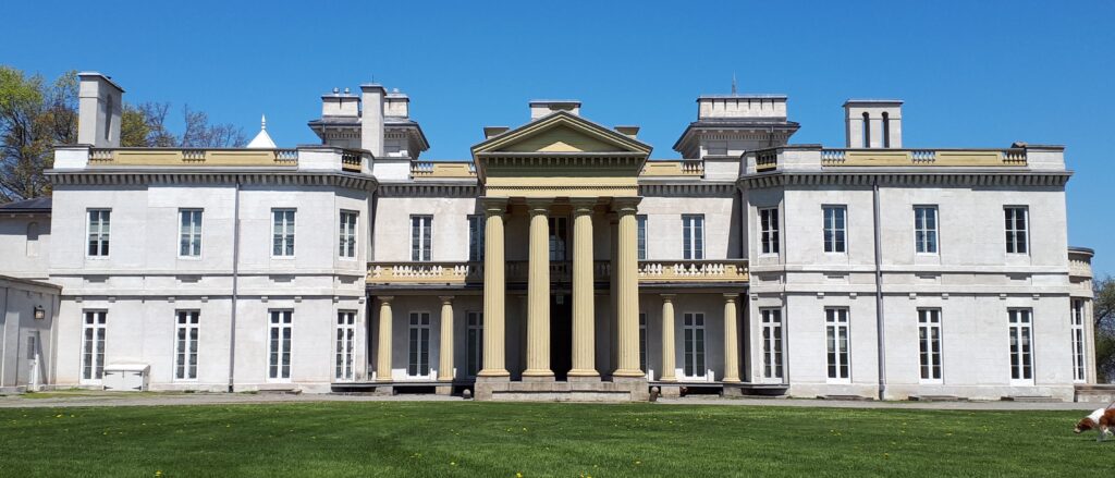 a large white house with four columns over the front door and wings extending out on either side. the house sits back on a green lawn against blue sky