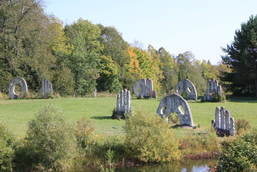 a large stone sculpture of a head with two eyes and two hands rise from the ground in a grass area that's surrounded by green and yellow trees