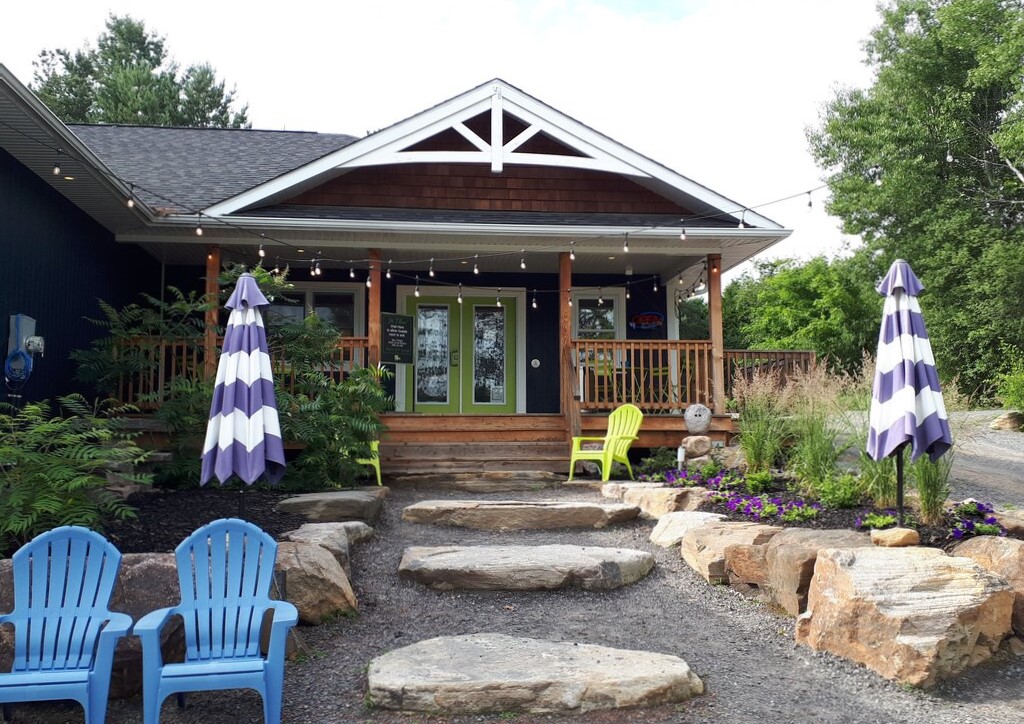 a wood building with strings of lights under the eaves faces a gravel path with two large stepping stones along the path. there's a yellow and blue muskoka chair along the path providing seating