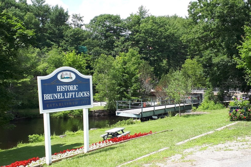 a sign stating 'historic brunel lift locks' stands above a bed of red and white flowers on a grass lawn beside the water.