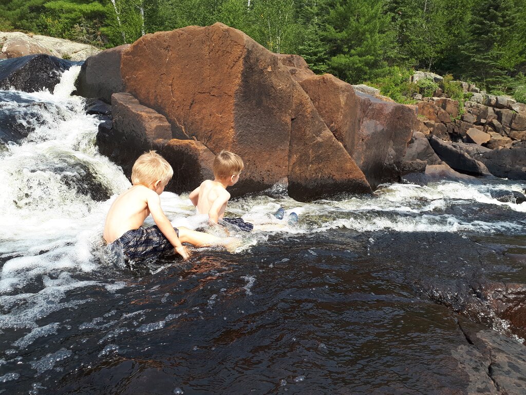 two boys sitting in a river as the water flows down beside two large brown rocks.