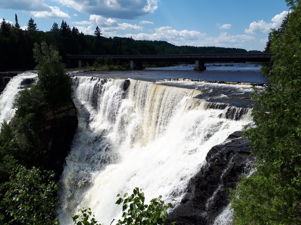 a large wide waterfall flowing over a high cliff. the view is looking at the top of the falls as the water tumbles over the edge. green trees surround the falls.