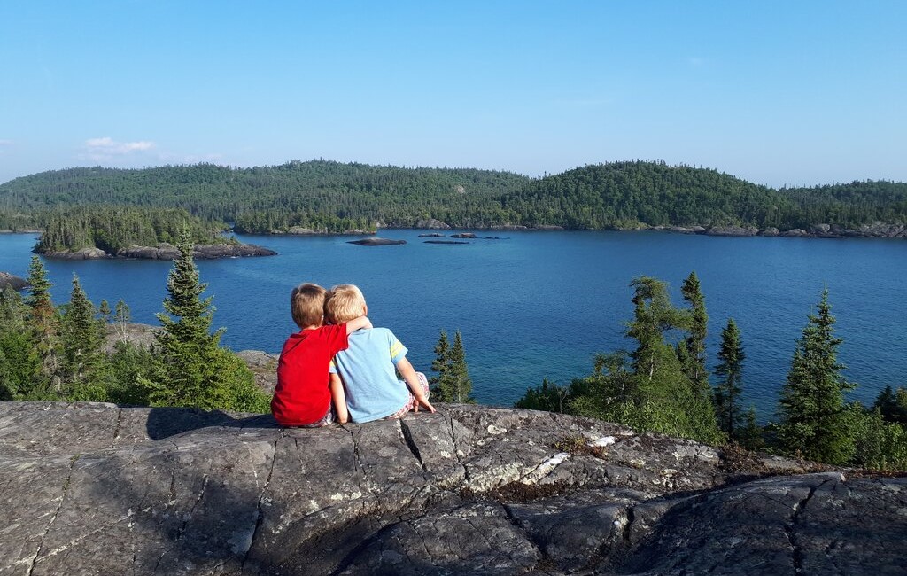 Two young children sit side by side on a rocky outcrop, gazing out over a calm, forested lake in Pukaskwa National Park, Ontario. Surrounded by evergreen trees and distant forested islands, the scene captures a peaceful, scenic moment in Canada’s rugged northern wilderness.