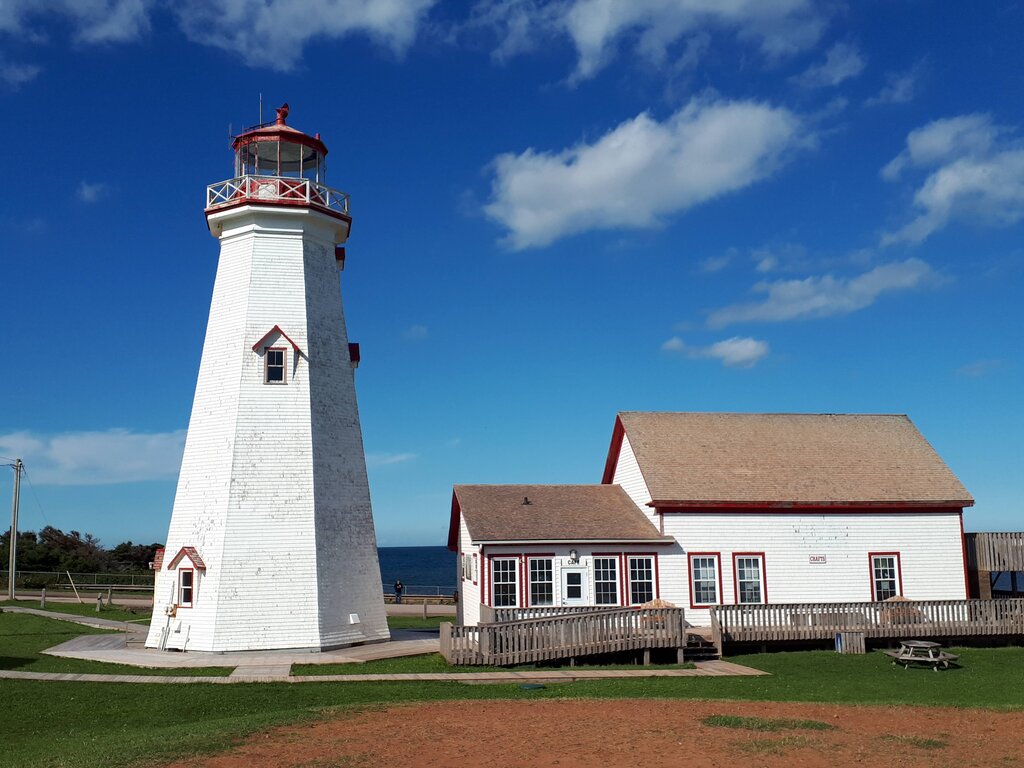 white lighthouse with red lantern room beside a house with attached room. The white house has a wood walkway and ramp and brown shingles