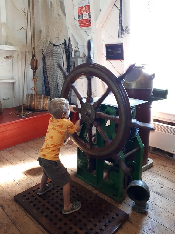 child in an orange shirt and shorts trying to turn the big ship wheel in the Souris lighthouse