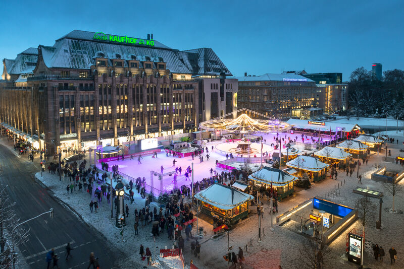 large skating rink under lights and surrounded by wooden vendor huts in front of a large building in Germany