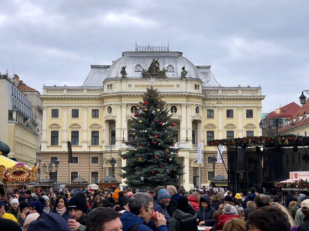 a large decorated Christmas tree sits in front of the Slovak National Theatre in Bratislava. people are milling about the Christmas market in front of the tree