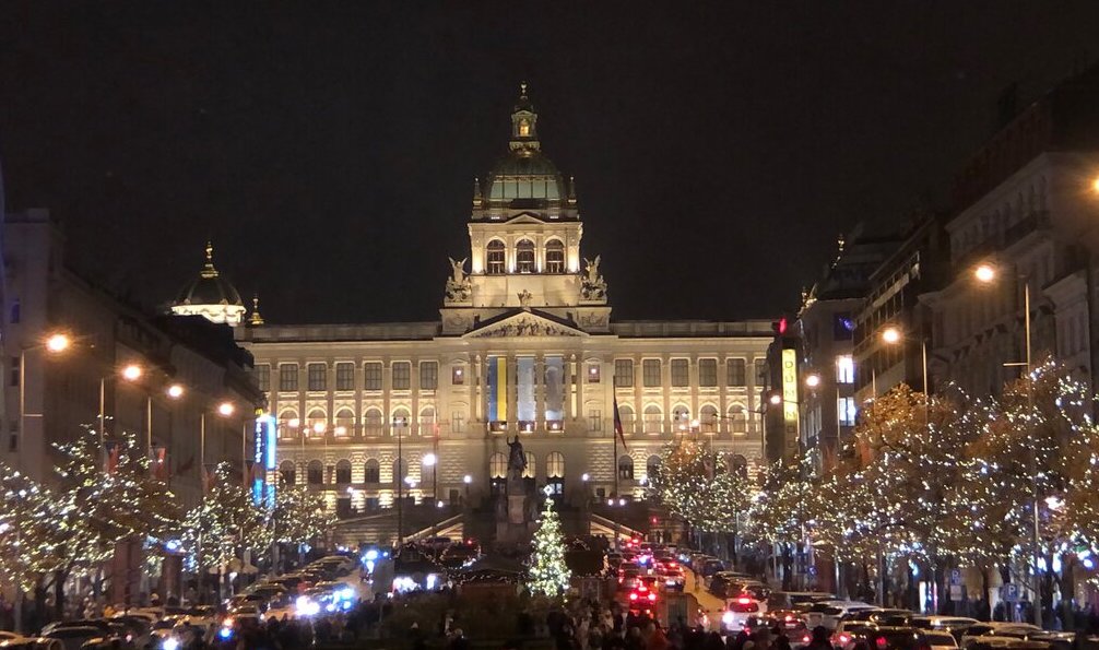 the national museum in prague is lit up at night and there is a lighted tree in front of the museum. trees along the square are lit up with lights