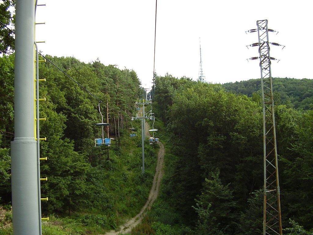 cable cars pass over a forested area where a narrow two lane path can be seen underneath. the Kamzik TV tower can be seen in the background