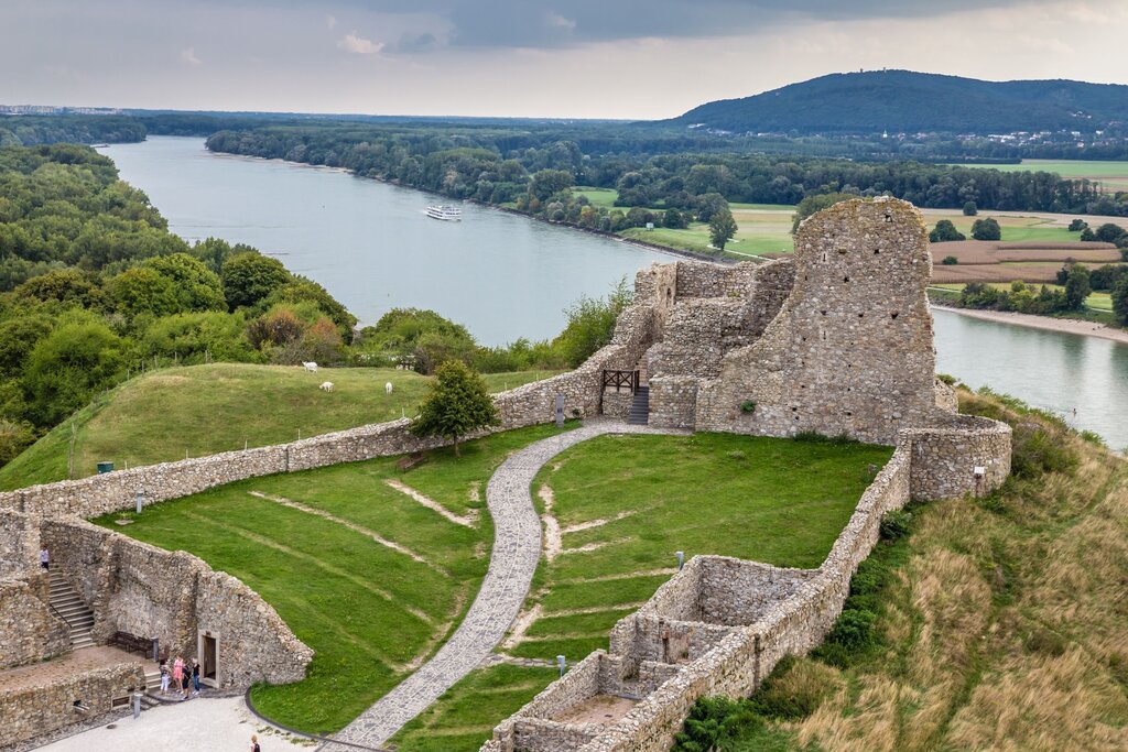 ruins of Devin castle stand on a cliff above the Danube River. walls can be seen and parts of the castle front near the edge of the river remain. a path leads through the inside of the castle to the remains at the river’s edge