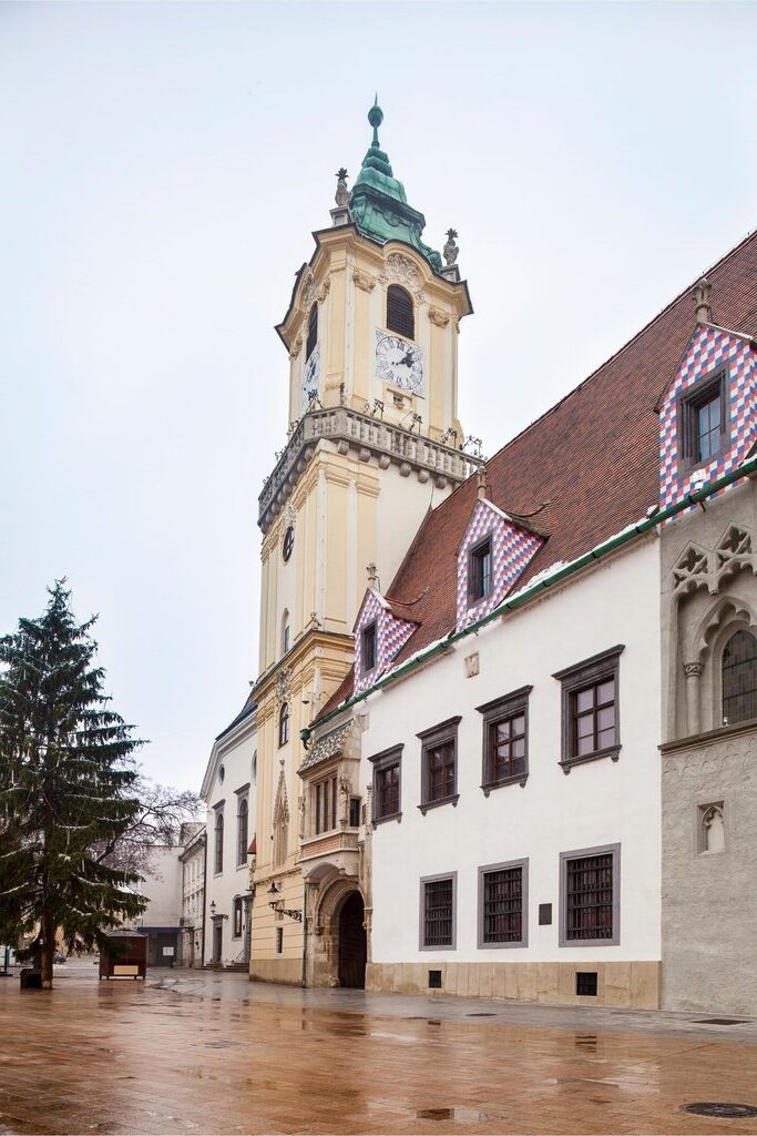 a tall tower sits amid a wall of houses and buildings in Bratislava. one building Is white with a red roof, beside it is a beige tower and gate at ground level, this is Michael’s gate, the only gate left in bratislava
