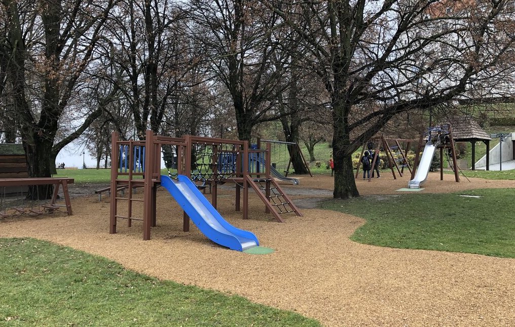 blue slide on a wooden climbing structure on a bed of wood chips. green grass surrounds the play area. a metal slide can be seen in the background on another wooden play area