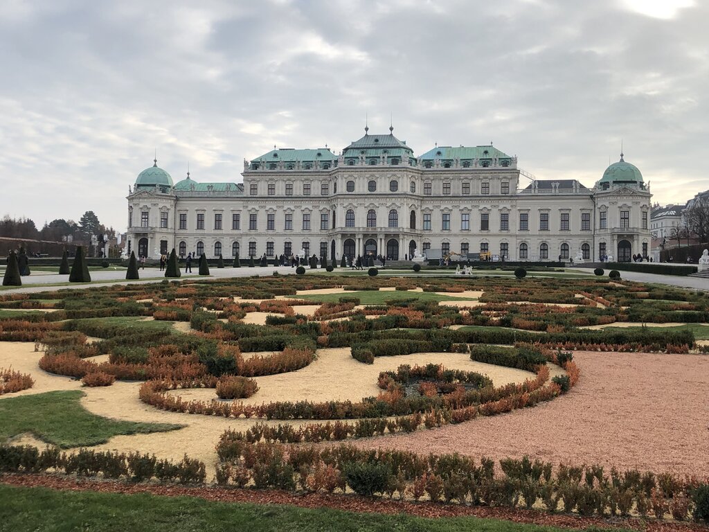 belvedere palace is a large white palace with many rectangular windows and a green roof. a large garden is in front with bushes shapes in circular designs