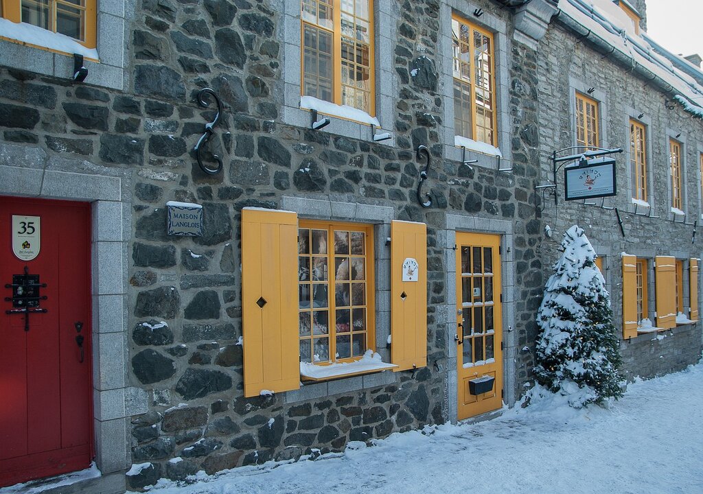 snow covered street with a stone building that has yellow shutters on the window and a yellow door in Old Quebec City in winter
