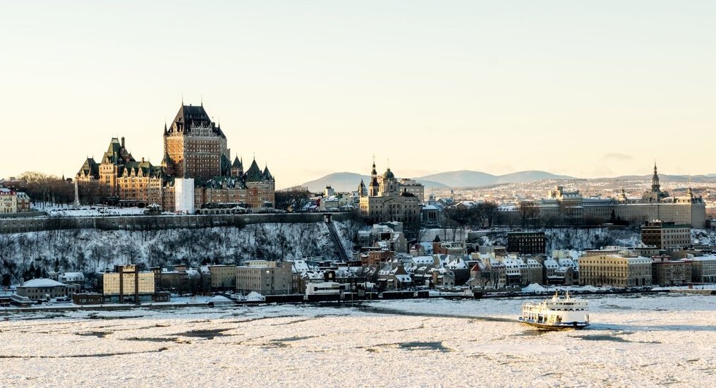 Chateau Frontenac sits high on the edge of the embankment overlooking the Saint Lawrence River while buildings can be seen along the water edge and a ferry cuts a path through the ice on the river