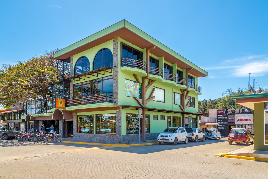 3 story green building on a corner with large arched windows on top floor, large rectangle windows on middle floor and a tree growing out of the window to the left side. cars are parked on the street in front of the right side of the building in Santa Elena, Monteverde