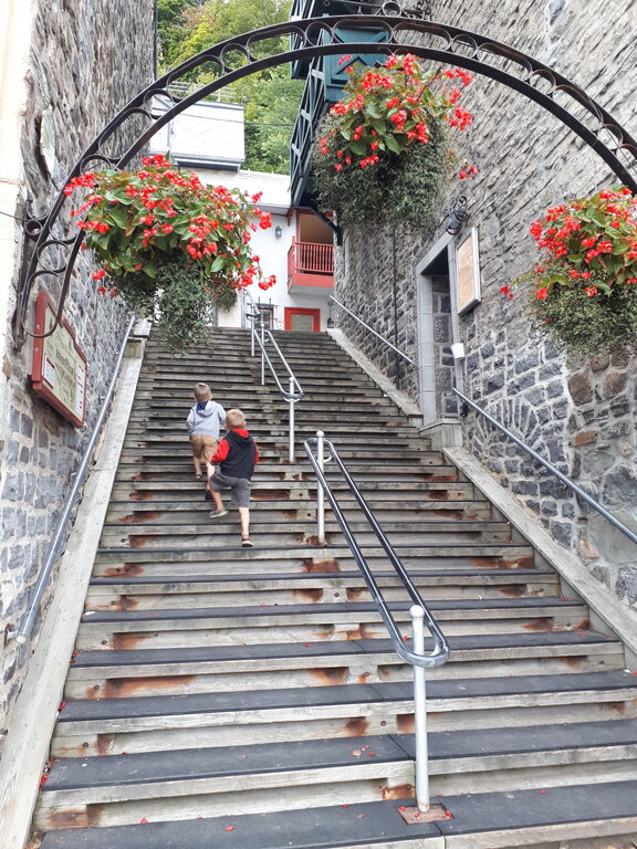 Two boys run up a charming stone stairway in Old Quebec, flanked by historic stone buildings and bright hanging flower baskets under an ornate black arch.