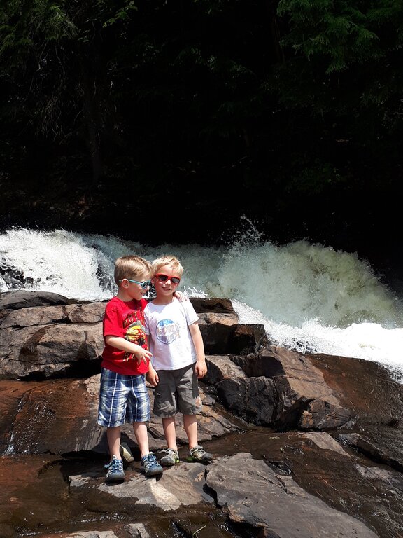 Two young boys wearing sunglasses pose on wet rocks in front of a rushing waterfall, surrounded by forest. One boy has his arm around the other as they smile in the sun.