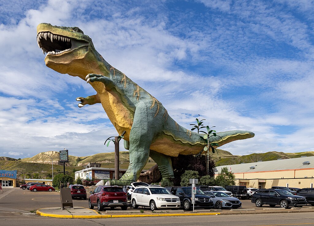 a huge t.rex dinosaur stands in the town of Drumheller, alberta, dwarfing the surroundings. four cars are right underneath the dinosaur's feet, reaching only a fraction up the leg of the dinosaur. the dinosaur's tail sits above the roof of a building to the right.