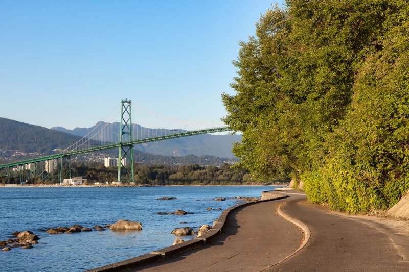 A scenic view of Vancouver’s Stanley Park Seawall with the Lions Gate Bridge in the distance, framed by ocean waters and lush green trees.