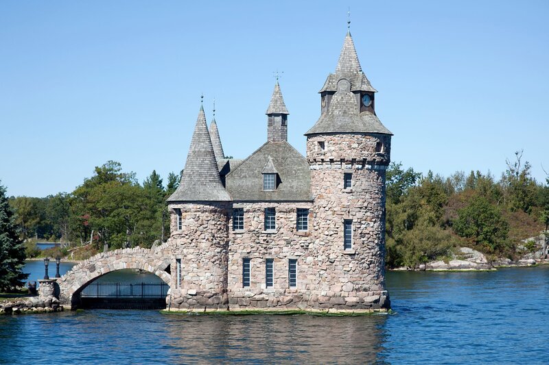 Stone castle with turrets and arched bridge sits on a small island surrounded by calm blue water, framed by trees under a clear sky. Boldt Castle in the Thousand Islands is a picturesque and historic gem, making it one of the most unique places to visit in Ontario.