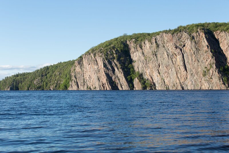 Sheer granite cliff known as Mazinaw Rock rises dramatically from the deep blue waters of Mazinaw Lake, lined with green trees along its crest. Located in Bon Echo Provincial Park, the rock face is famous for its Indigenous pictographs and striking natural beauty.