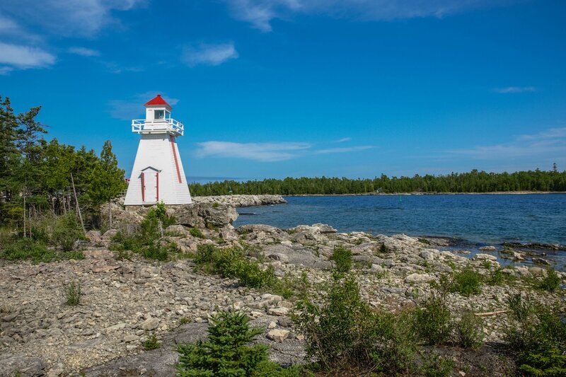a short white lighthouse sits on a rocky beach next to the water in manitoulin island