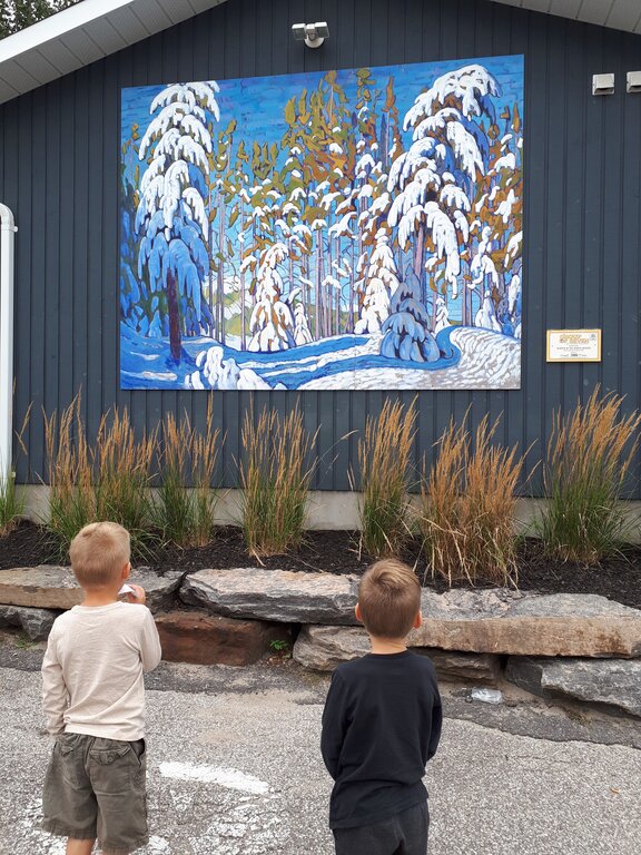 Two boys look at a large mural showing a snow-covered forest with tall pine trees under a bright blue sky. The mural is mounted on a dark exterior wall of a building, with decorative grasses planted below it.