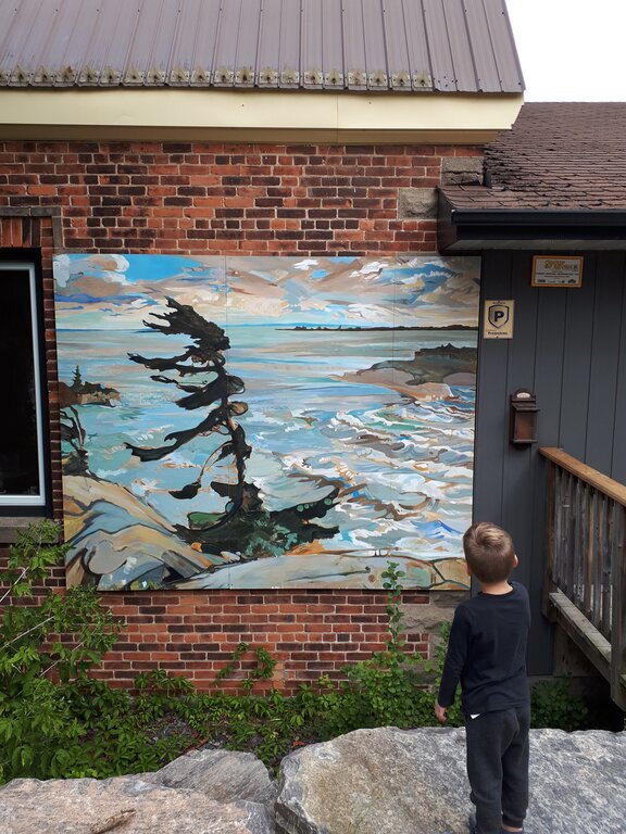 A child stands on a stone ledge gazing at a mural of a windswept pine tree overlooking a rocky shoreline and waves, painted on a brick wall. The mural, a replica of a Group of Seven painting, uses expressive brushstrokes to capture a coastal landscape under a moody sky.