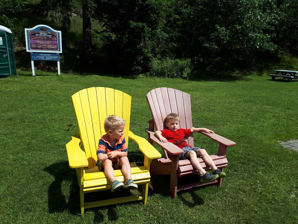 Two boys relax in oversized wooden Adirondack chairs on a grassy lawn. One chair is painted bright yellow and the other a faded red, with a park sign with history of the Brunel Lift Locks in Huntsville stands in front of trees in the background.