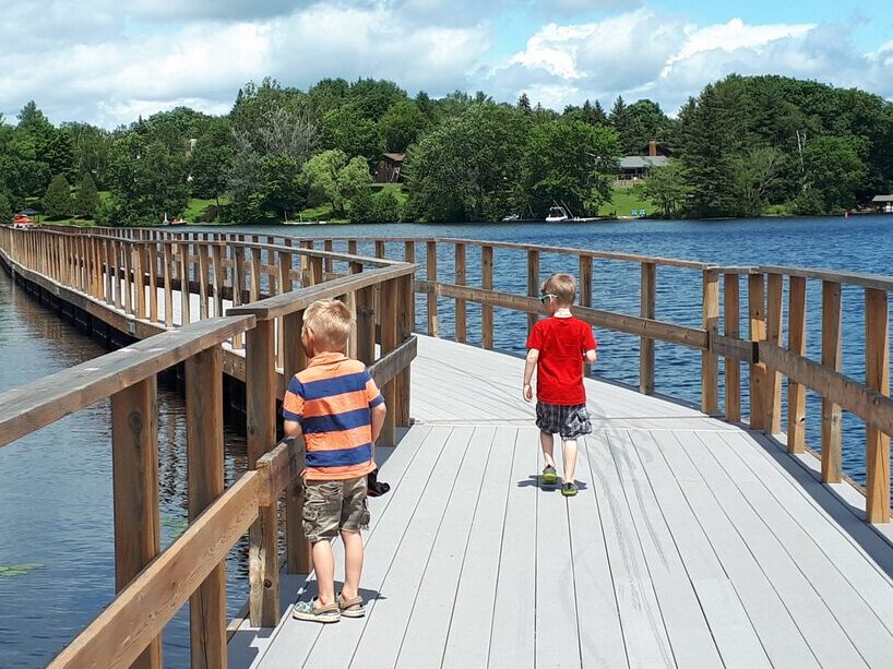 Two boys walk along a wide wooden boardwalk curving over a lake, with trees and houses visible on the far shore. Puffy clouds float in a partly sunny sky.