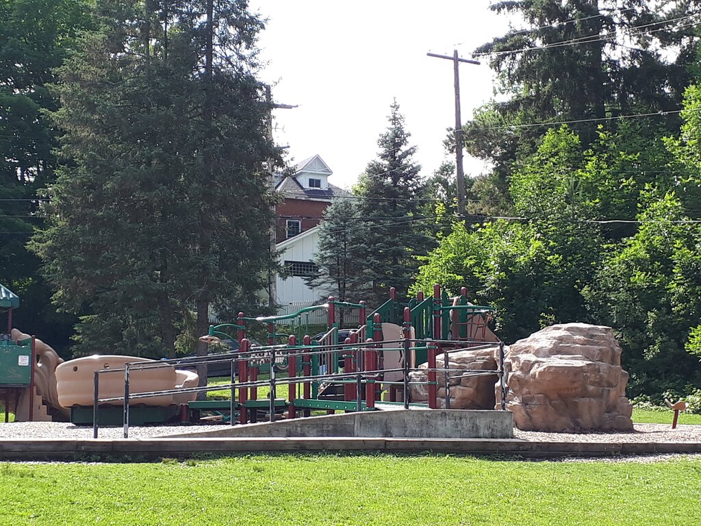 A playground with tan and green equipment is surrounded by grass, large faux boulders, and tall trees. A white house peeks through the trees in the background.