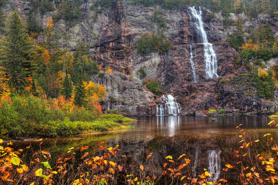 Bridal Veil Falls cascades down a rugged cliffside in Agawa Canyon, framed by colorful autumn foliage and reflected in the still water below. The multi-tiered waterfall flows dramatically over dark rock, surrounded by dense forest in vibrant reds, oranges, and greens.
