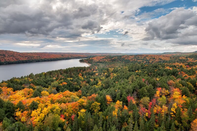 A panoramic view from a trail in Algonquin Park, showcasing a vibrant autumn forest with a mix of fiery red, orange, and yellow foliage among evergreens. A winding lake cuts through the dense trees under a dramatic sky with layered clouds and patches of blue.