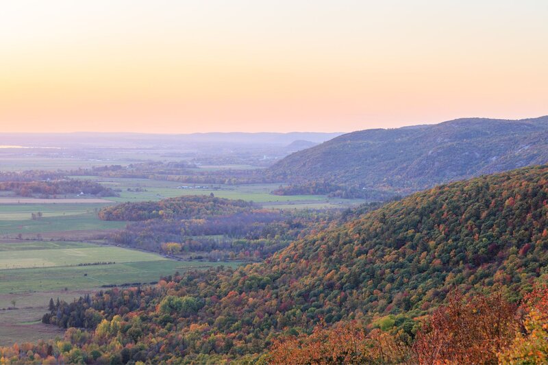 A soft, pastel-toned sunset view from Champlain Lookout in Gatineau Park, overlooking a sweeping valley dotted with early autumn foliage. Rolling hills and open farmland stretch into the distance under a calm, hazy sky.