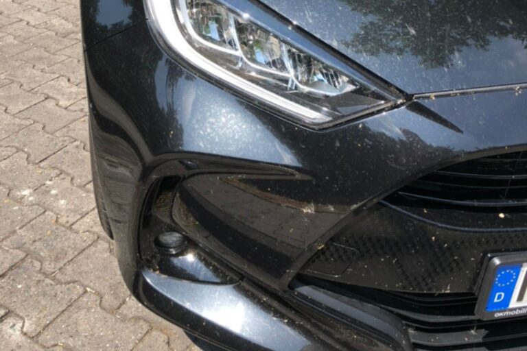 Close-up of the front left corner of a dark-colored car, showing a sleek headlight design, glossy bumper with a lower air intake, and a European-style license plate partially in view. The car surface is speckled with dirt and debris, suggesting recent use.