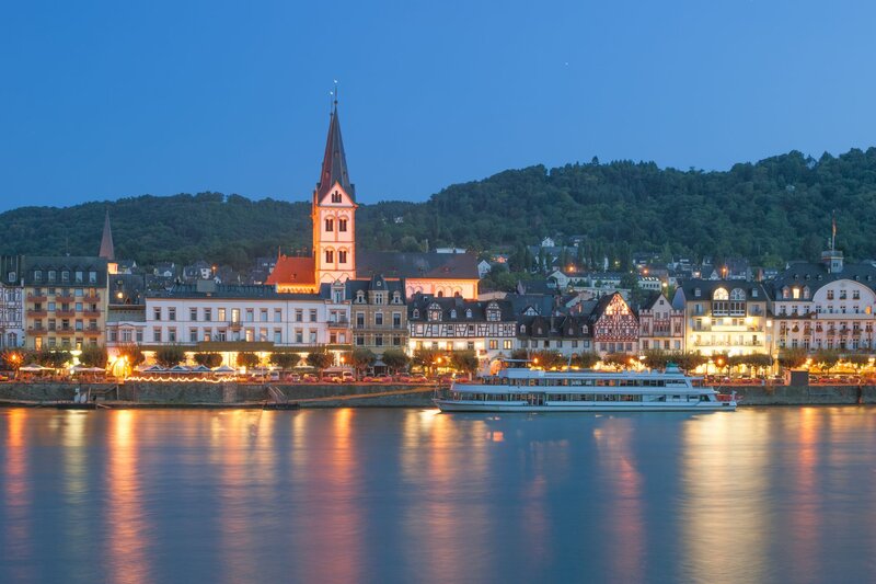 Evening view of Bacharach, Germany, with its illuminated half-timbered buildings and church tower reflecting on the calm waters of the Rhine River. A river cruise boat is docked along the lively waterfront, while forested hills form a peaceful backdrop under a twilight sky.