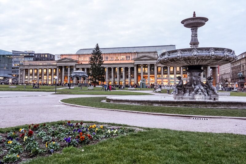 A wide-angle view of Schlossplatz in Stuttgart, Germany, featuring the grand Königsbau building with glowing interior lights, a large stone fountain with sculpted figures in the foreground, and neatly kept flower beds and walkways throughout the square. A tall Christmas tree stands near the building, adding seasonal charm.