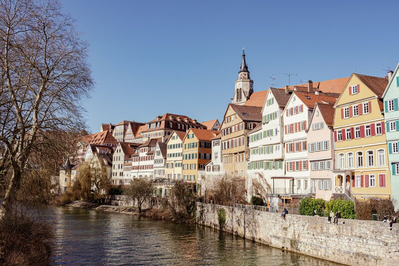 A row of colorful half-timbered houses lines the Neckar River in Tübingen, Germany, under a clear blue sky. The pastel buildings with steep gabled roofs create a charming, historic atmosphere, while a stone embankment and sparse winter trees frame the riverside walkway.