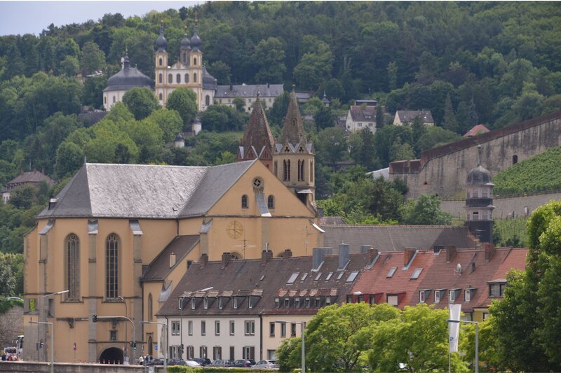 A scenic view of Würzburg, Germany, showcasing a large tan church with twin pointed towers and a golden clock on its facade, set against a backdrop of terraced vineyards and forested hills. Higher up on the hill, the twin-domed pilgrimage church Käppele stands out among scattered houses and lush greenery.