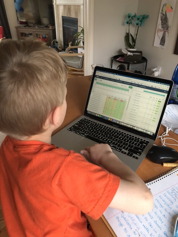 A boy in an orange shirt works on a laptop displaying a colourful spreadsheet with rankings and data. A notebook with handwritten notes lies open beside him on the table